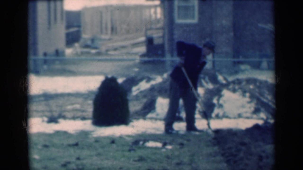 A man pauses over his shovel in a snowy Michigan yard, framed by a dark shrub, fence, and nearby residential buildings.