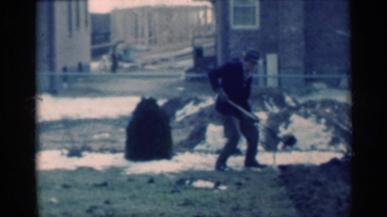 The man crouches and pulls the shovel through exposed dirt, with patchy snow, a shrub, fence, and residential buildings in view.