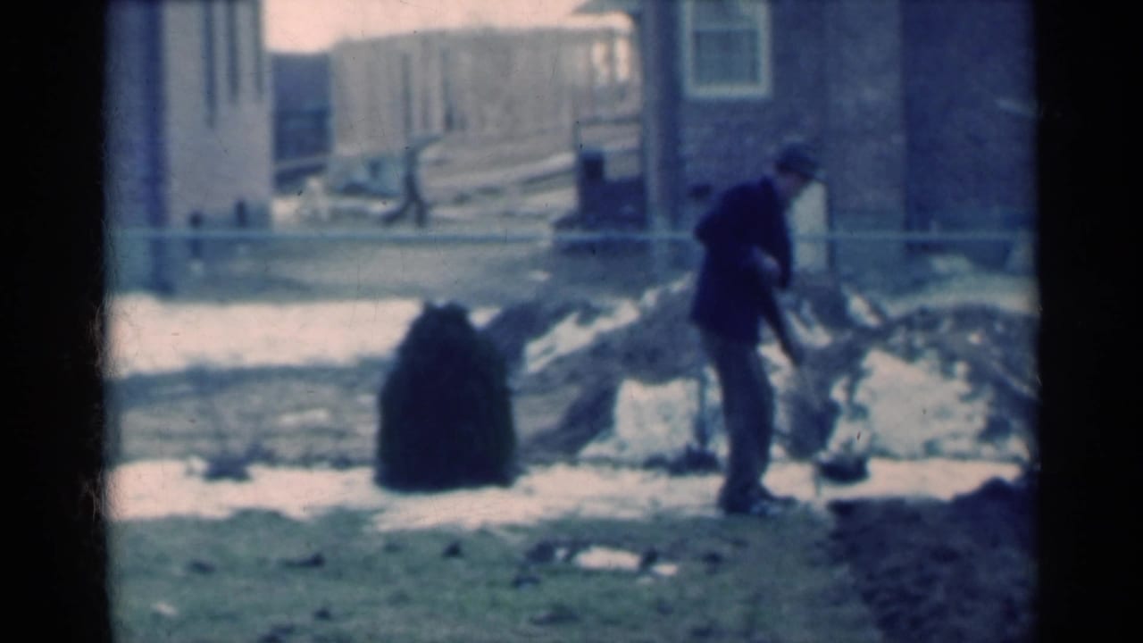 Man in a dark suit and hat shoveling near snow patches in a residential yard.