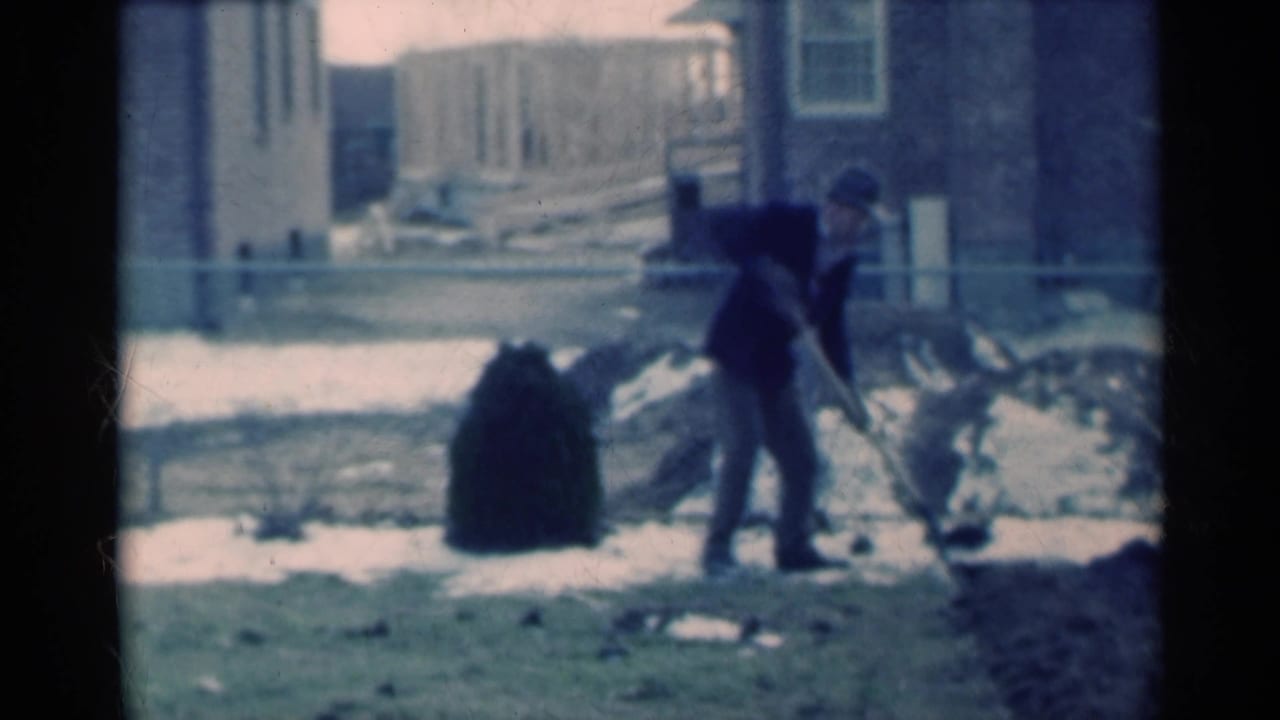 A man bends while using a shovel in a 1939 Michigan residential yard, with snow, bare ground, and house exteriors visible.