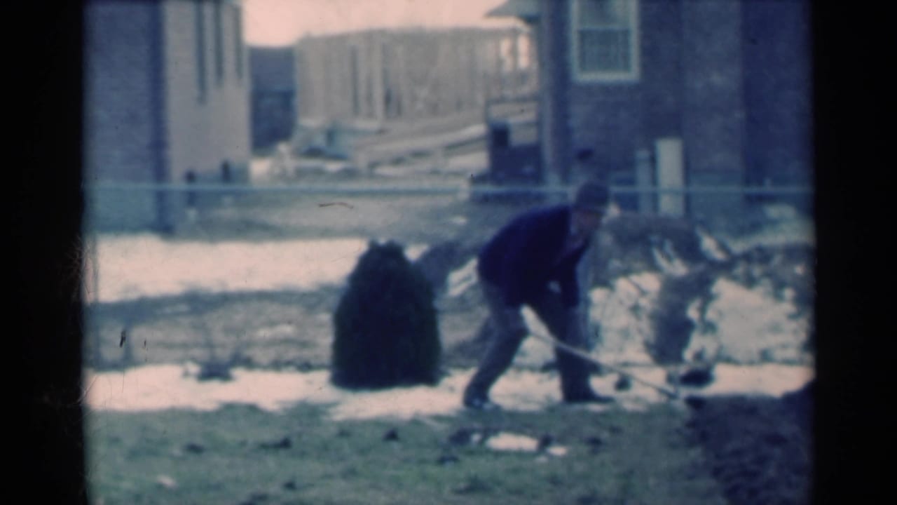 The man continues shoveling at the edge of the Michigan yard, framed by snow patches, bare soil, a shrub, and residential buildings.