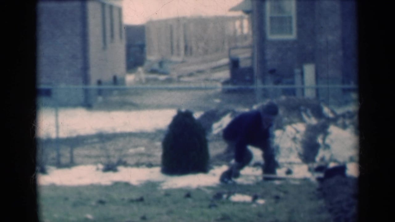 Man shoveling in a snowy yard beside brick houses and a chain-link fence.