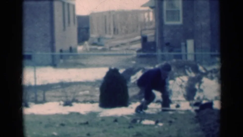 In 1939 Michigan home-movie footage, a man bends with a shovel in a snow-patched residential yard near brick houses and a fence.