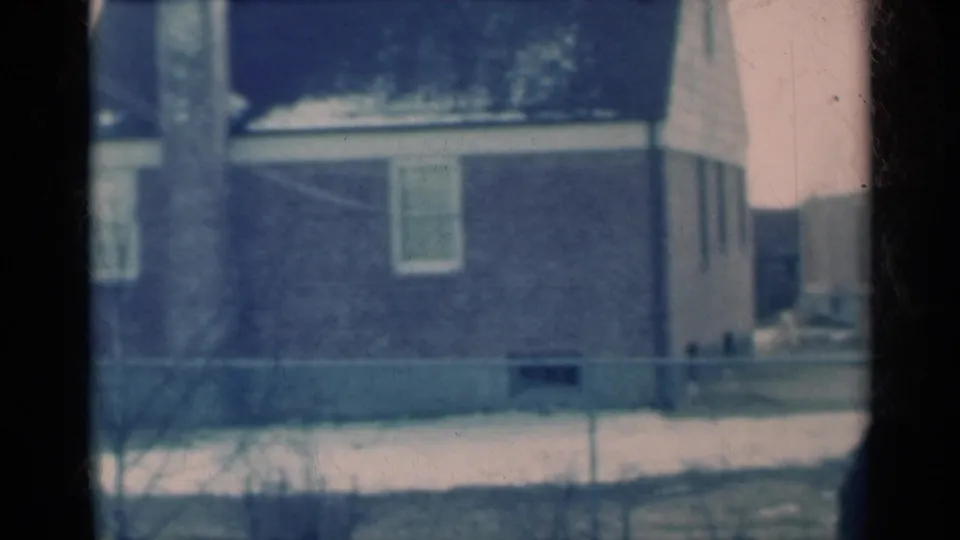 The camera frames a brick residential house, chain-link fence, bare shrubs, and patches of snow in a Michigan neighborhood.