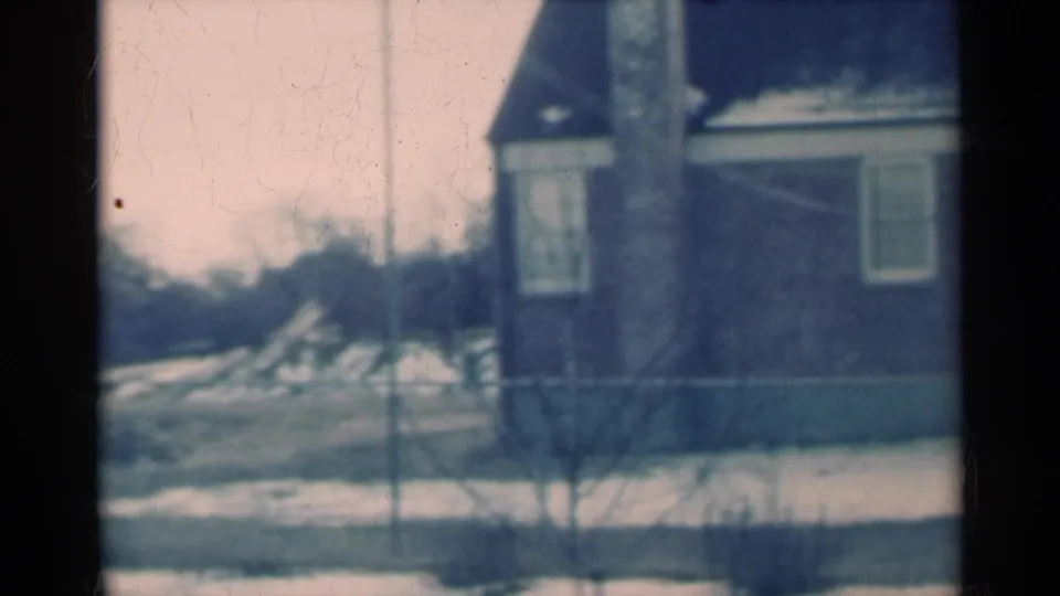 A winter residential view shows a brick house, chain-link fence, bare tree trunk, and snow-patched yard in Michigan.
