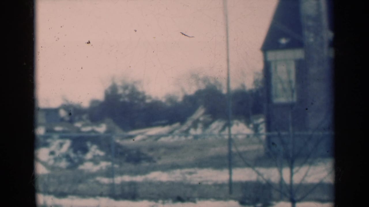 Snowy rural view through a dark window frame with a small tower-like building and bare trees.