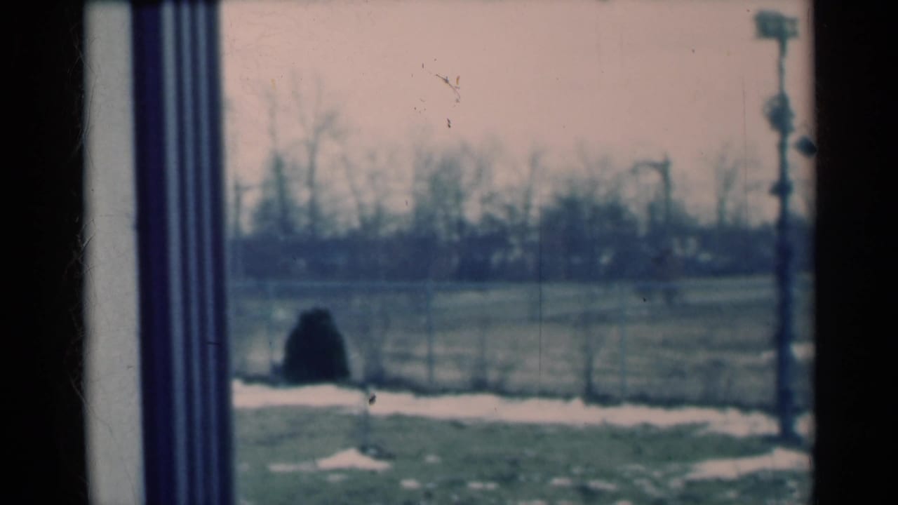 1939 Michigan home-movie frame showing a winter yard through a window, with fence line, bare trees, snow patches and utility pole equipment.