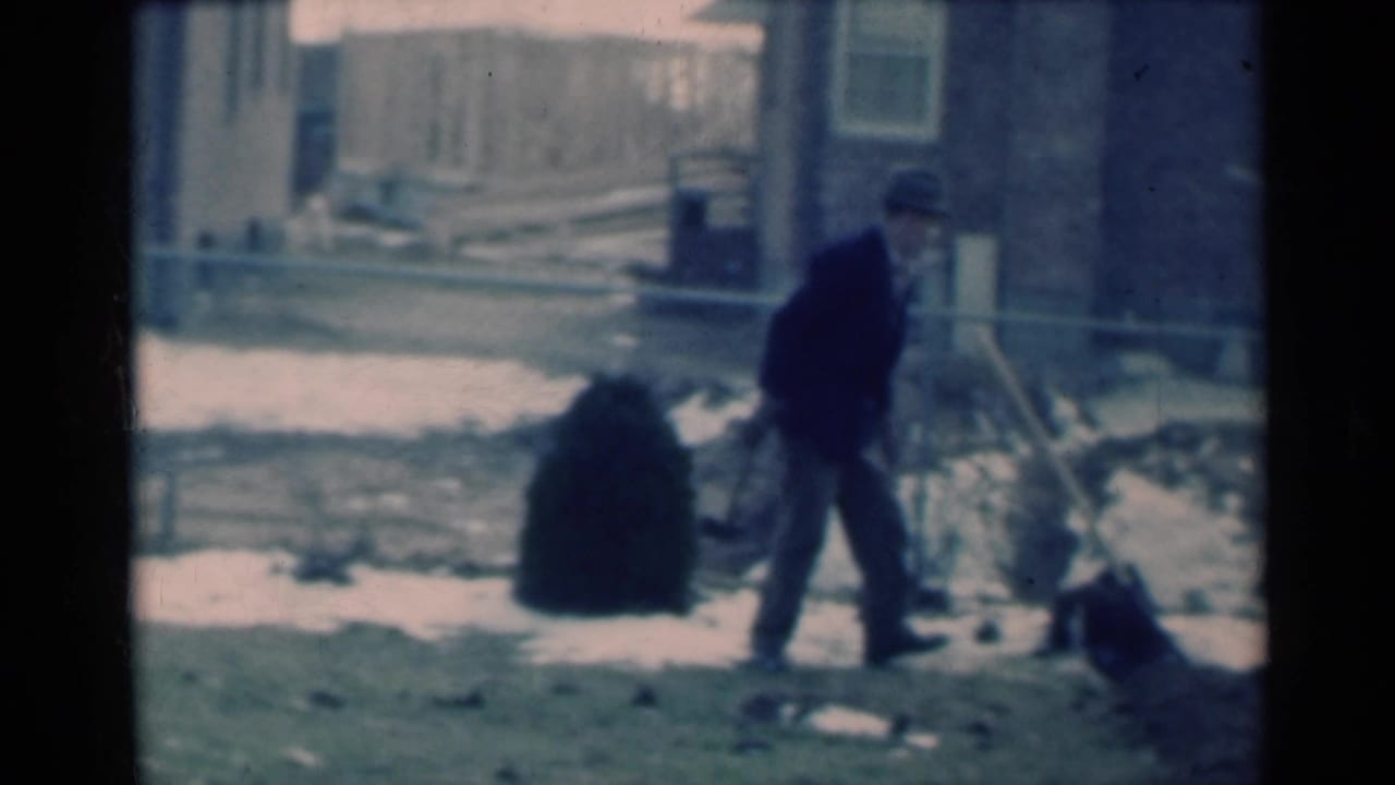 Michigan 1939 frame with an adult man in dark winter clothing working in snow beside a brick house.
