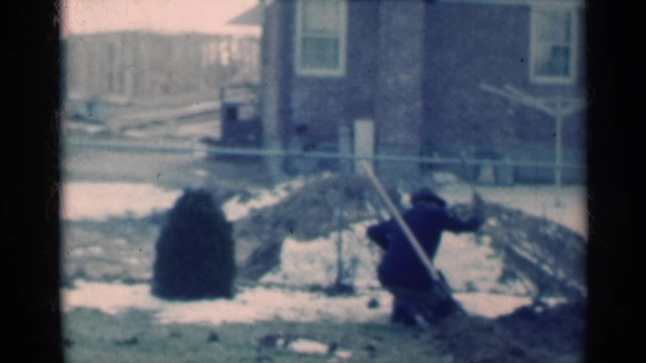 Michigan 1939 frame with the man crouched in a snowy yard, continuing to break or dig into frozen ground near the house.