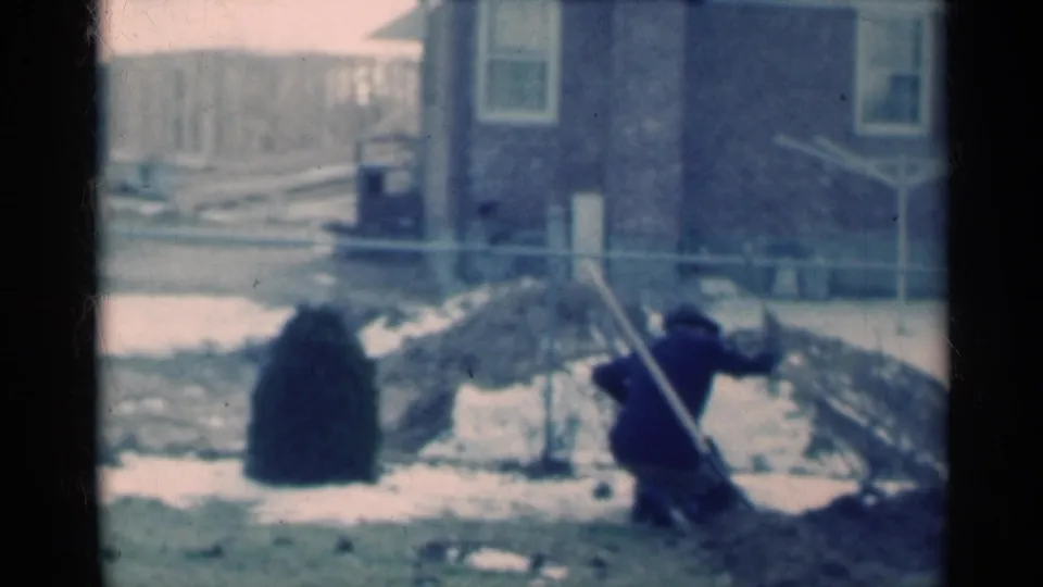 Michigan 1939 frame with the man crouched in a snowy yard, continuing to break or dig into frozen ground near the house.