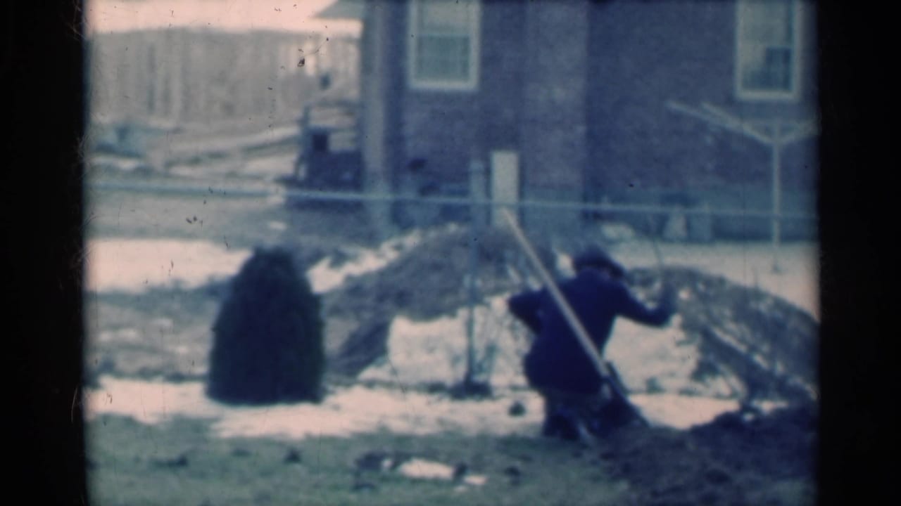 In 1939 Michigan home-movie footage, a man in dark clothing crouches in a fenced backyard with snow patches, dirt mounds, and a house behind him.