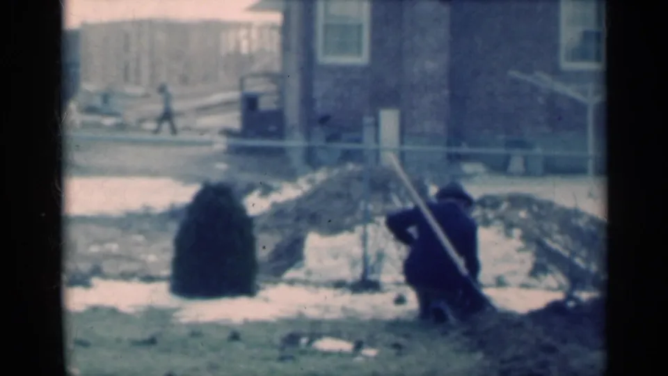A man continues yard work in a Michigan backyard, with a distant passerby, fence, snow patches, dirt piles, and house exterior visible.