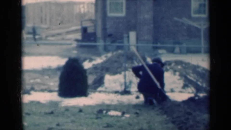 The 8mm frame shows a man crouched beside a leaning stake in a Michigan backyard, surrounded by snow patches, dirt mounds, and residential fencing.