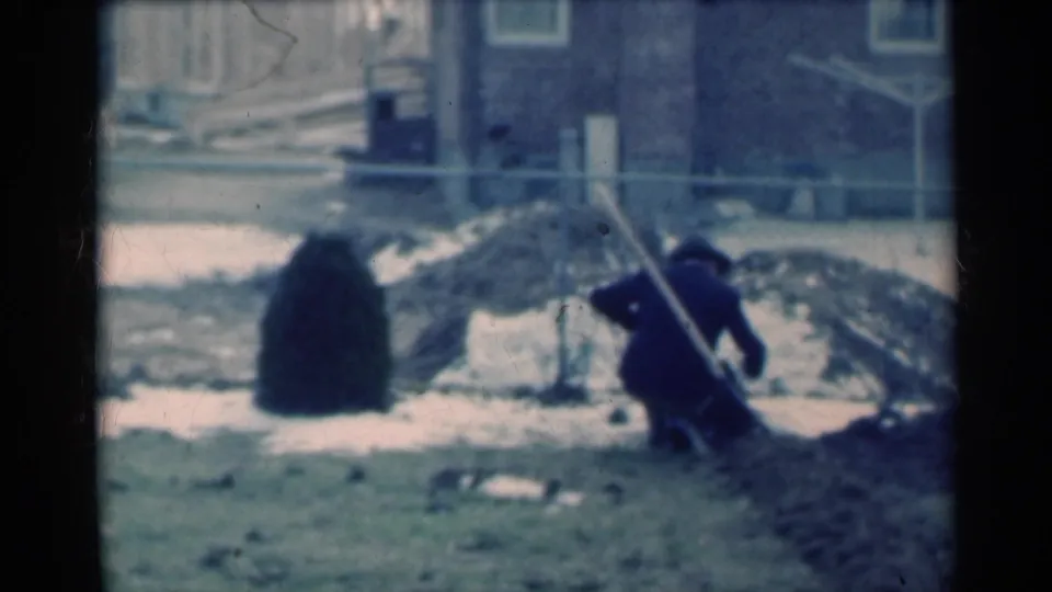 A man in dark clothing leans over backyard soil near a stake, with snow-dappled ground, a small evergreen, fencing, and a house behind him.