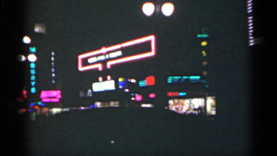1949 New Orleans night-street frame with bright neon signage, lit storefront windows, dark sidewalk figures, and visible 8mm frame edge.
