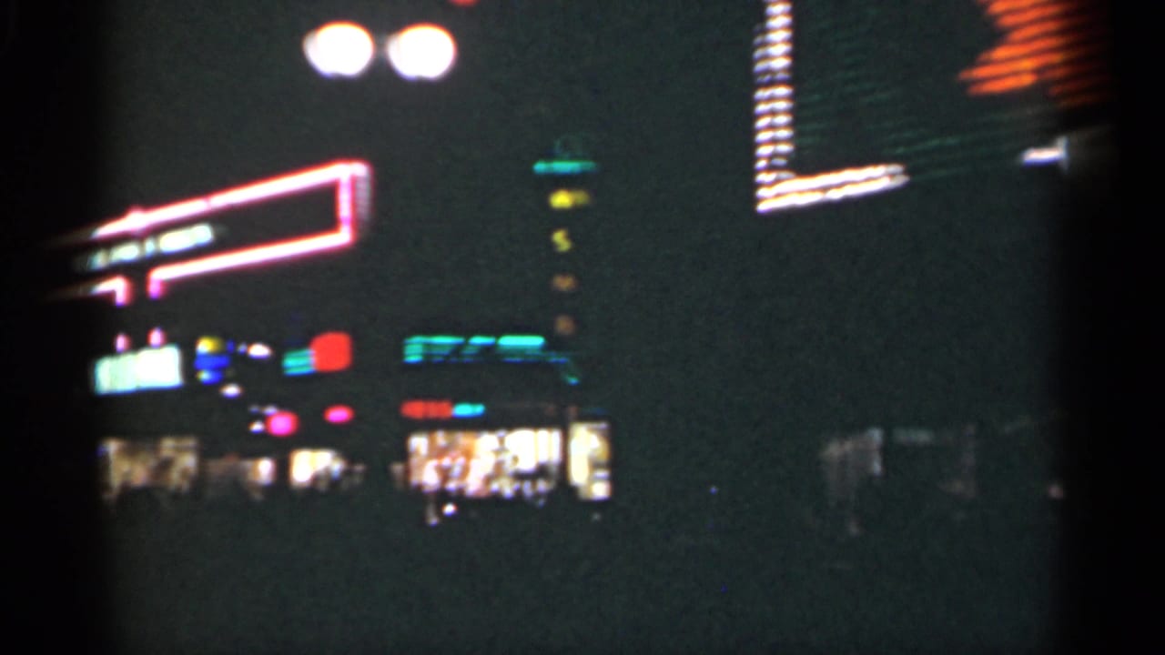 Dark nighttime street view with blurred illuminated storefront signs and colored lights.