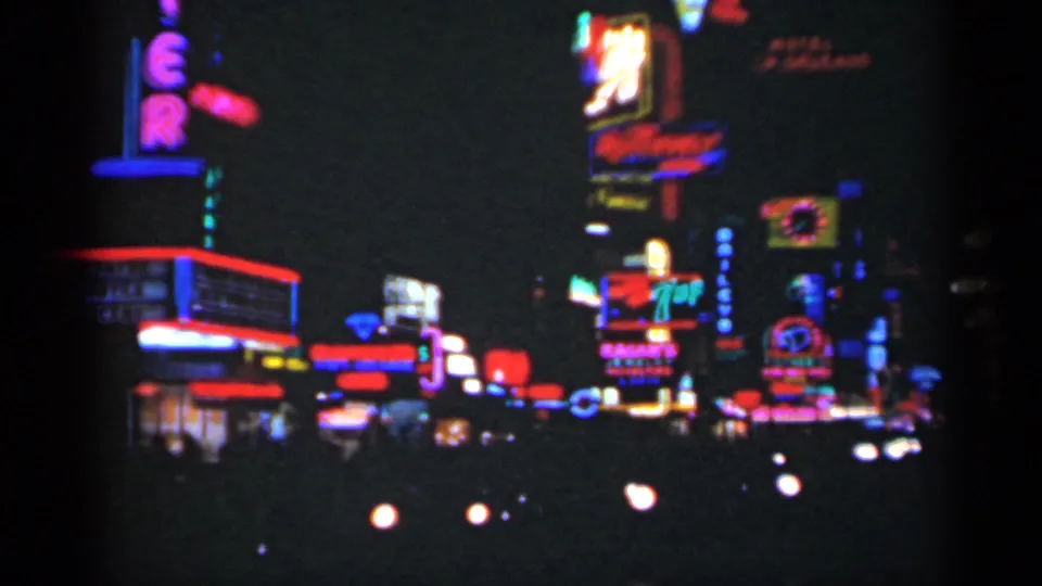 1949 New Orleans nighttime street view with bright neon advertising, dark roadway, and approaching headlights in soft 8mm focus.