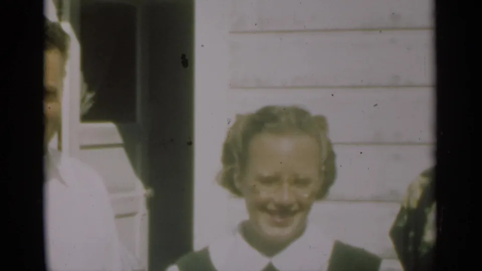 A woman with styled hair and glasses smiles toward the camera outside a white clapboard residence in New Orleans footage from 1949.