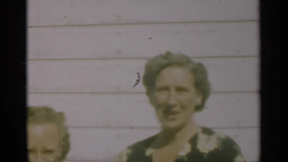 A woman in a dark floral dress stands outside against white clapboard siding in the 1949 New Orleans home-movie footage.