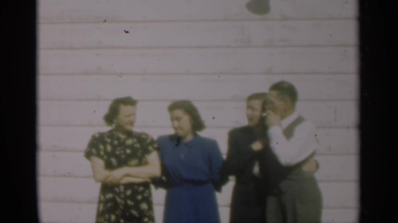 In the 1949 New Orleans home-movie frame, four adults pose casually outside a pale clapboard house, one man partly obscured.