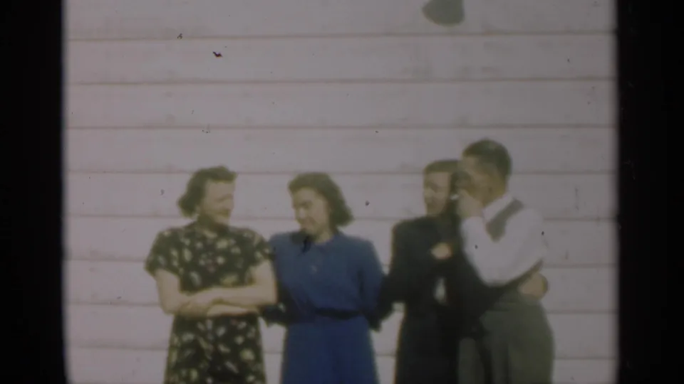 In the 1949 New Orleans home-movie frame, four adults pose casually outside a pale clapboard house, one man partly obscured.