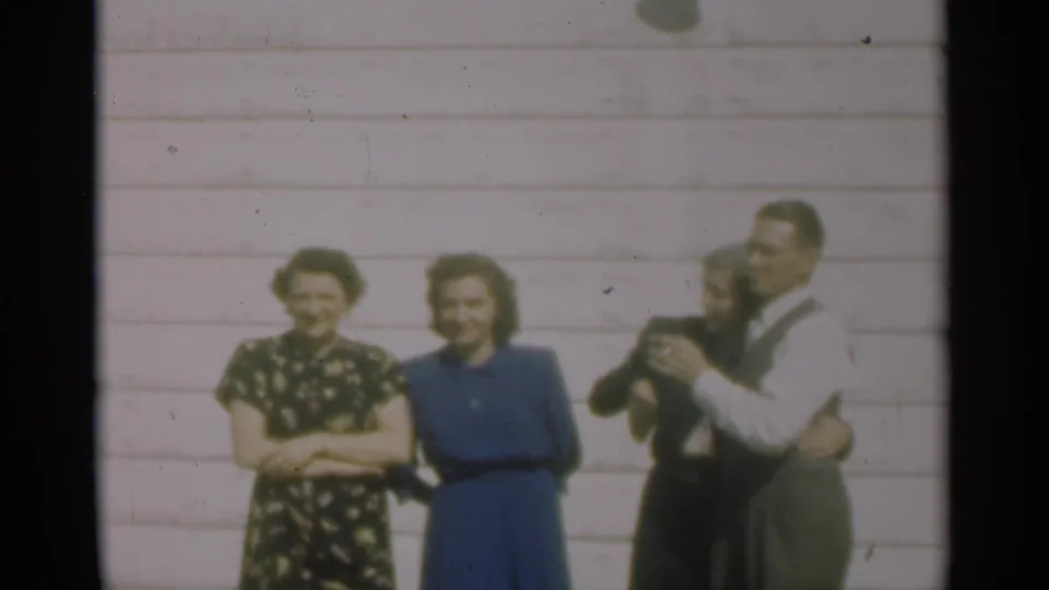 A 1949 New Orleans frame shows adults smiling by a clapboard wall while a man at right raises a cigarette.