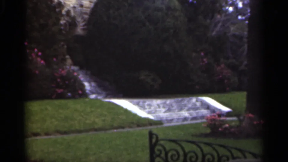 A distant person walks through a 1949 New Orleans garden near shrubs, stone steps, lawn, and decorative iron railing.