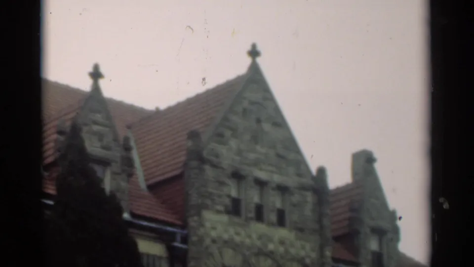 1949 New Orleans frame of a large stone exterior with steep gables, red roof tiles, dark evergreen foliage, and pale overcast sky.