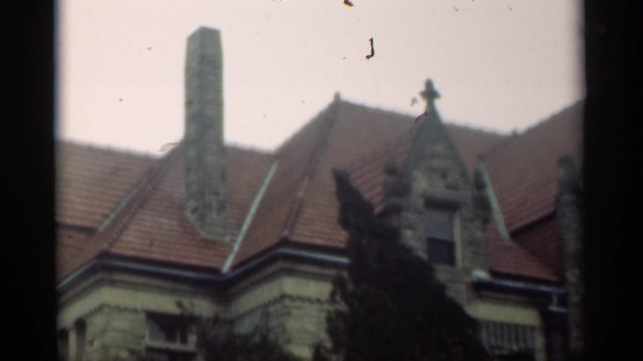 Stone house roofline with tall chimney, red tiles, gable details, and trees in front.