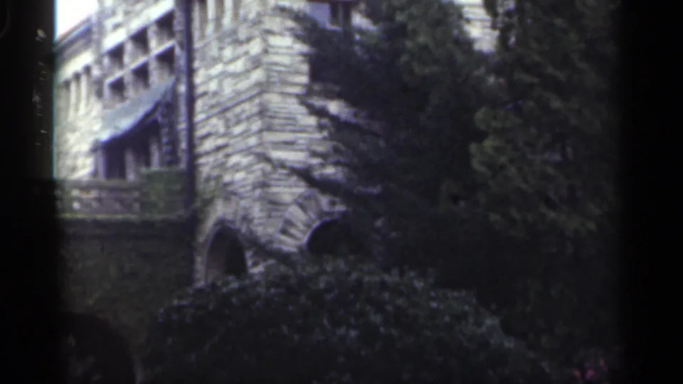 1949 New Orleans 8mm frame of the stone house wall, partly hidden by trees and shrubs, with a small lower arch visible.