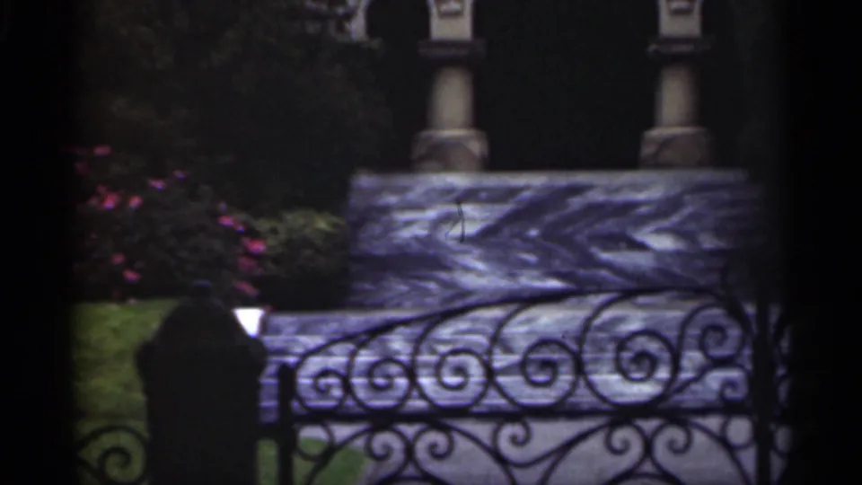 1949 New Orleans 8mm frame showing a wrought-iron fence, carved or textured stone steps, columns, garden flowers, and shaded greenery.