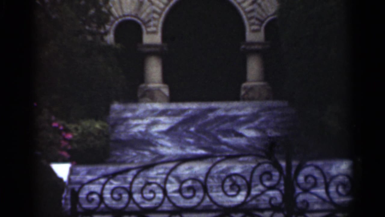 Ornate stone facade with arched openings, columns, railing, greenery, and a small lamp.