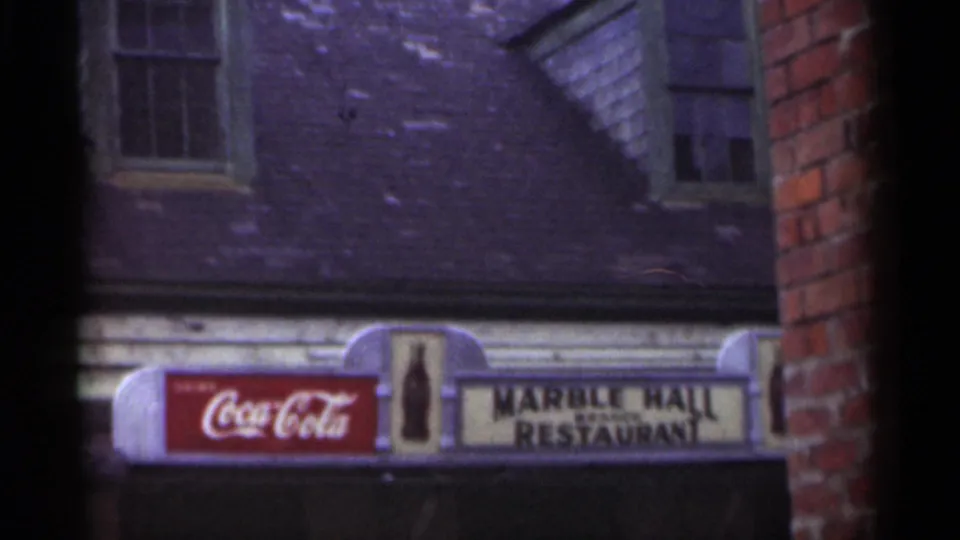 1949 New Orleans frame showing the Marble Hall Restaurant sign, a red Coca-Cola panel, dormer windows, brick edges, and gray roof shingles.