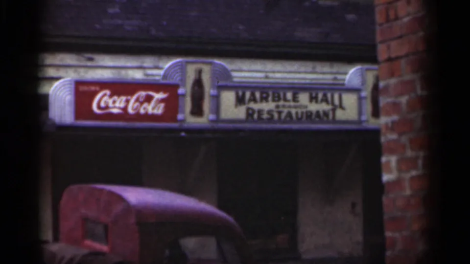 1949 New Orleans street-level frame of the Marble Hall Restaurant and Coca-Cola signs, with brick framing and a red vehicle beneath.