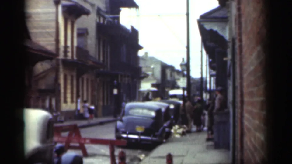 1949 New Orleans street view widening to parked cars, pedestrians, building facades, and brick framing in soft 8mm color.