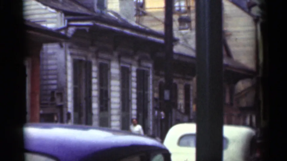 New Orleans street in 1949 with older building fronts, parked automobiles, a utility pole, and a distant person on the sidewalk.
