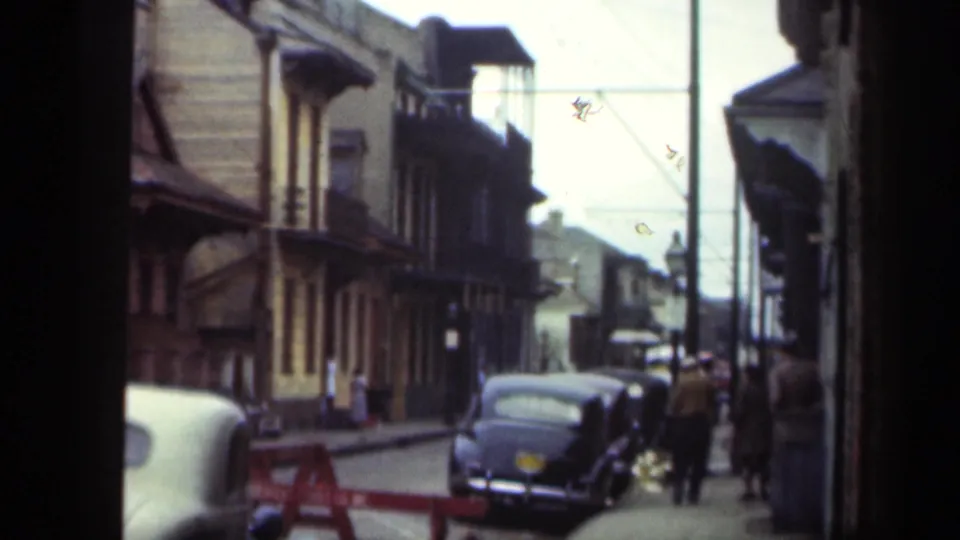 1949 New Orleans sidewalk and street view with parked cars, older facades, utility poles, and several pedestrians gathered or walking near the curb.