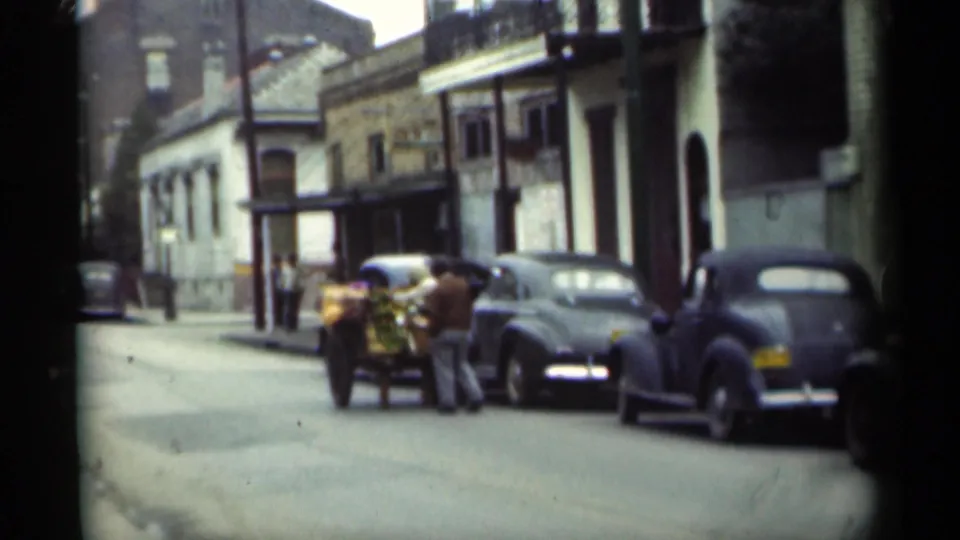 A New Orleans street in 1949 with a cart of goods, classic automobiles, pedestrians, and mixed brick and stucco building fronts.