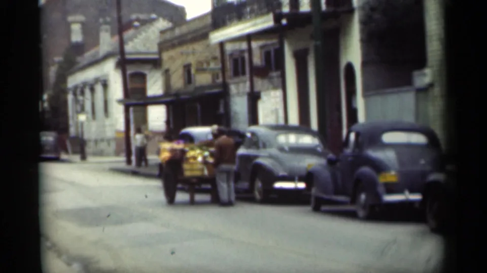 1949 New Orleans curbside view with pedestrians around a loaded cart, parked classic cars, utility poles, and street-facing buildings.