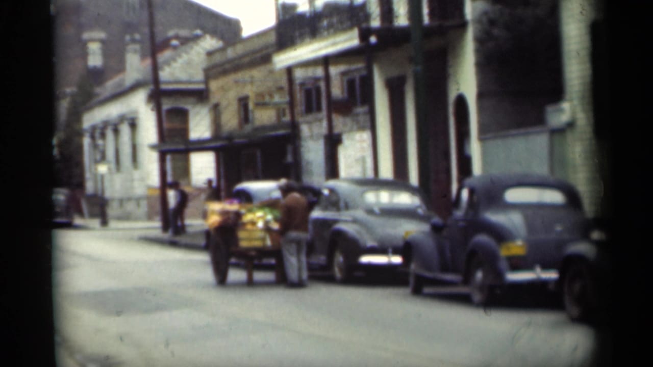 Street with parked classic cars, pedestrians, and a cart near older buildings.