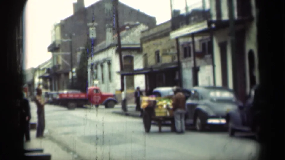 Wider 1949 New Orleans street frame with a red truck, parked cars, pedestrians, and a cart in front of older buildings.