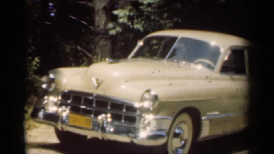 Close 1949-era view of a cream-colored classic sedan with chrome trim, yellow plate, and leafy background.