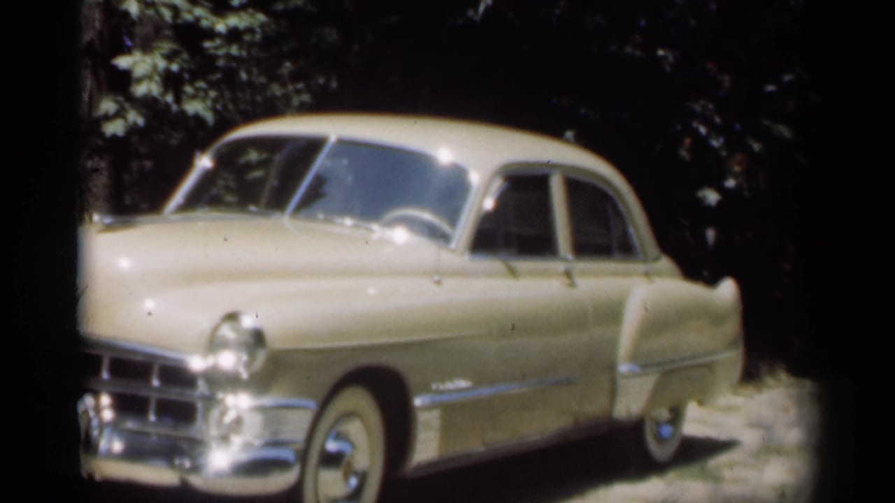 Cream-colored vintage sedan parked by trees with chrome bumper and whitewall tire visible.