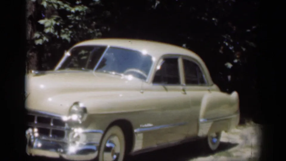Cream late-1940s sedan in a 1949 New Orleans 8mm frame, with chrome trim, whitewall tire, and shaded trees behind.