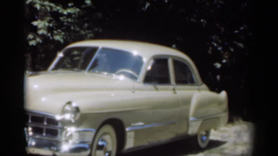 1949 New Orleans frame showing a cream vintage sedan parked off-road, with rounded fenders, chrome details, and trees nearby.