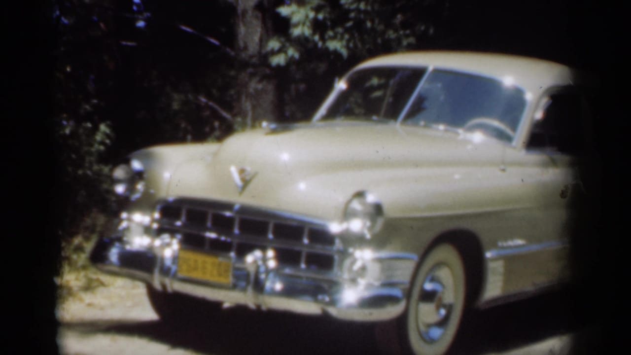 Beige vintage sedan parked on a dirt path under trees, seen from the front left.