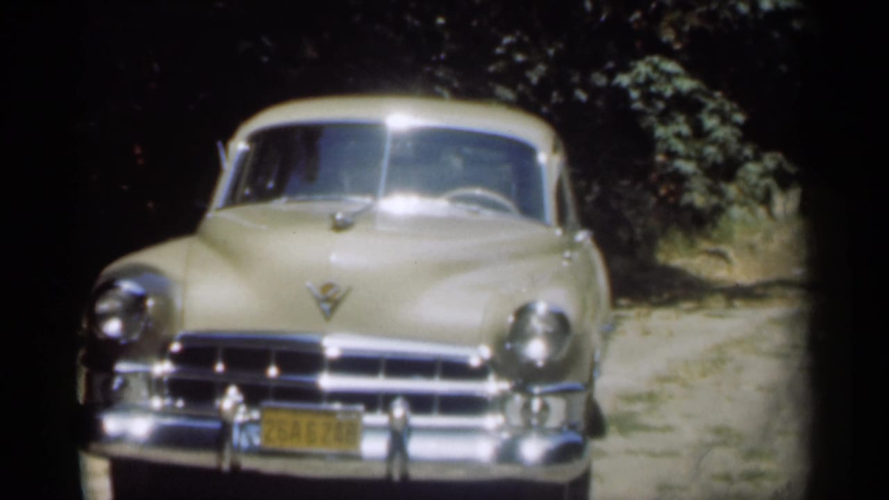 Cream Cadillac facing camera on a shaded dirt road with foliage behind it.