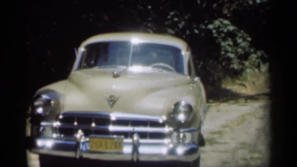 Front view of the cream Cadillac, likely from the 1949 New Orleans home-movie clip metadata, with foliage and dirt road surrounding it.