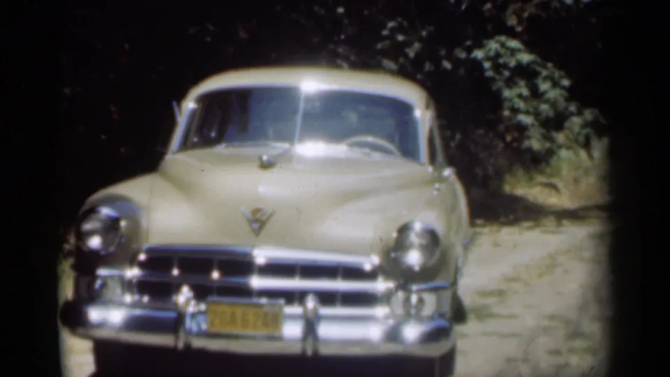 Archival 8mm frame of a cream Cadillac viewed head-on, with its chrome front end, yellow plate, and shaded roadside setting visible.