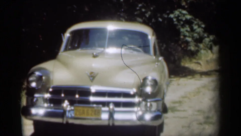 Final frame of the cream Cadillac on the wooded dirt road, with a dark film scratch crossing the windshield and hood area.